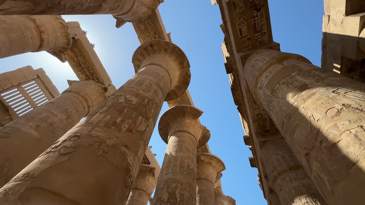 Una ripresa cinematografica dal basso che guarda verso l'alto le massicce colonne di pietra con capitelli a papiro e antichi geroglifici nella Grande Sala Ipostila del Tempio di Karnak.