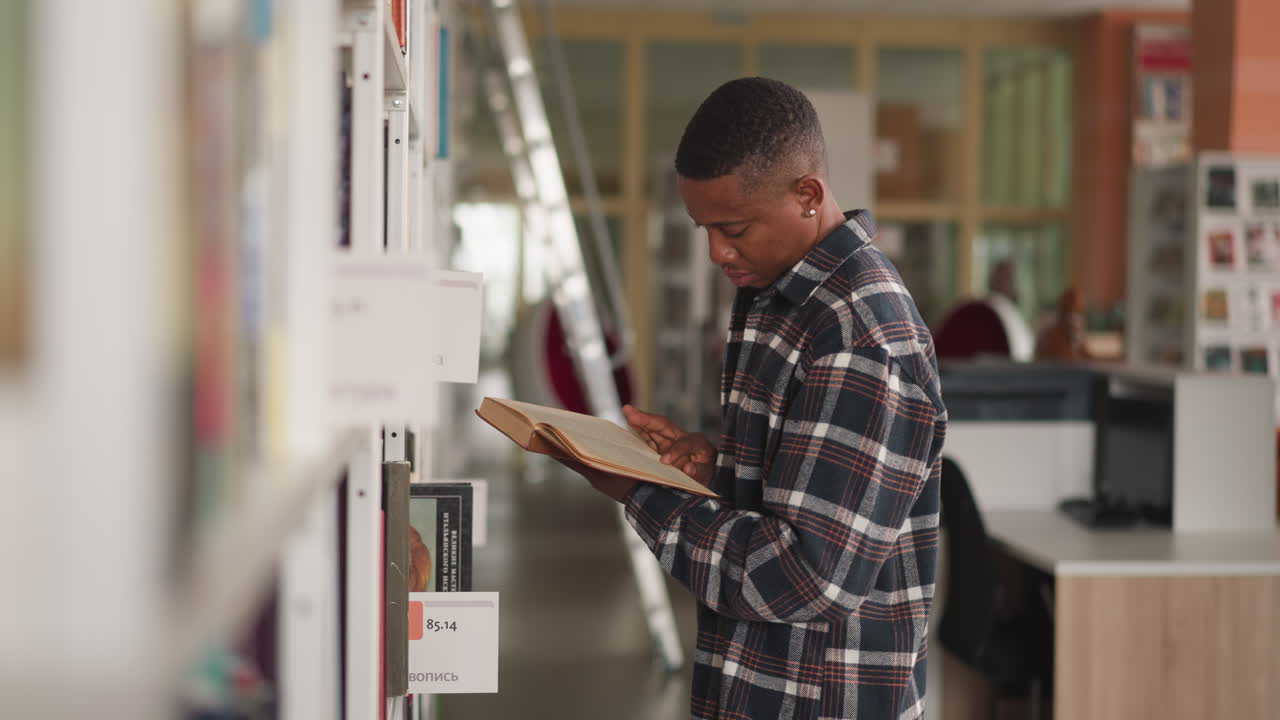 un hombre de piel oscura examina un libro cerca de una estantería. un estudiante afroamericano estudia el libro con fluidez antes de recogerlo para leer en casa. un tipo lee una novela.