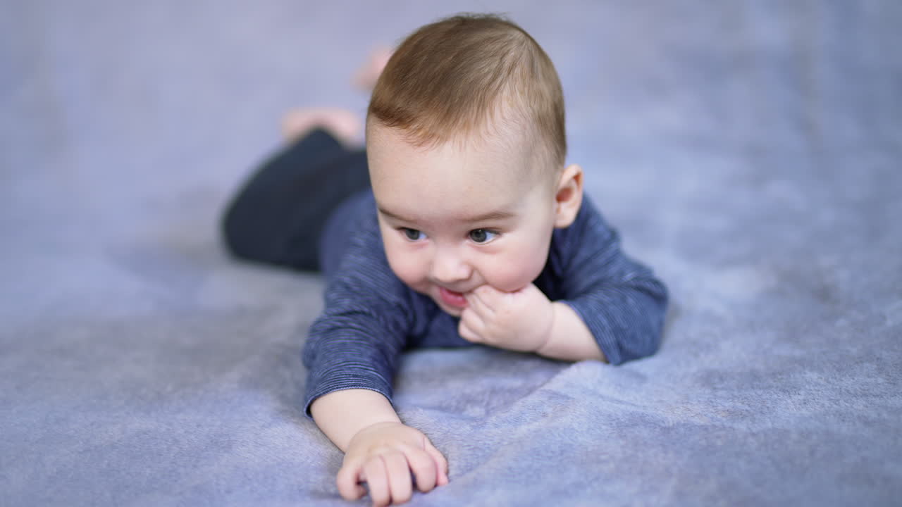 Adorable healthy child lies on bed and puts his fingers into mouth. Adult's hand plays with a kid trying to cheer him up.