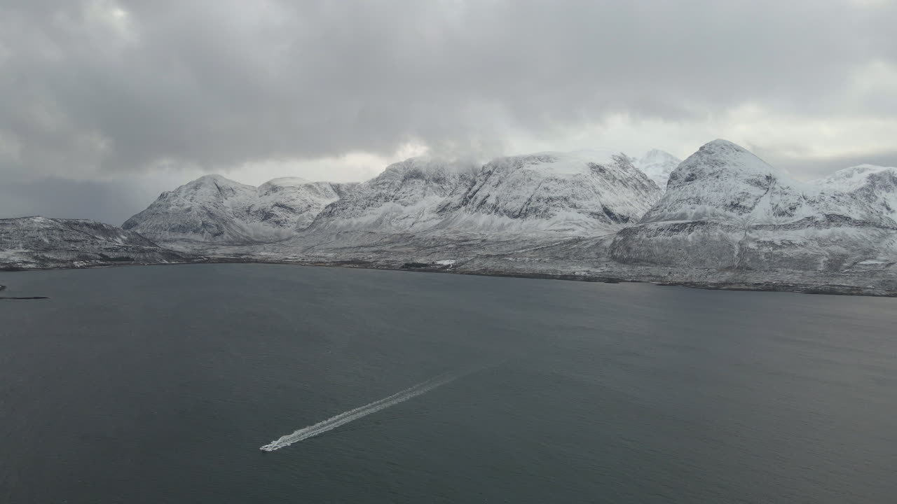 Drönarbild från hög höjd över en båt som snabbt färdas över ett lugnt hav, med snötäckta berg i bakgrunden