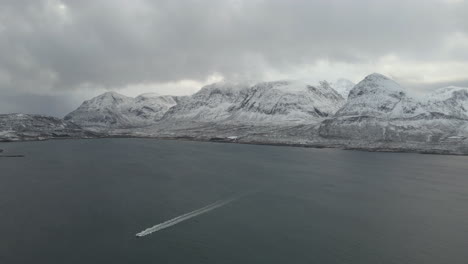 High-angle-drone-view-of-boat-speeding-along-calm-ocean,-mountains-covered-in-snow