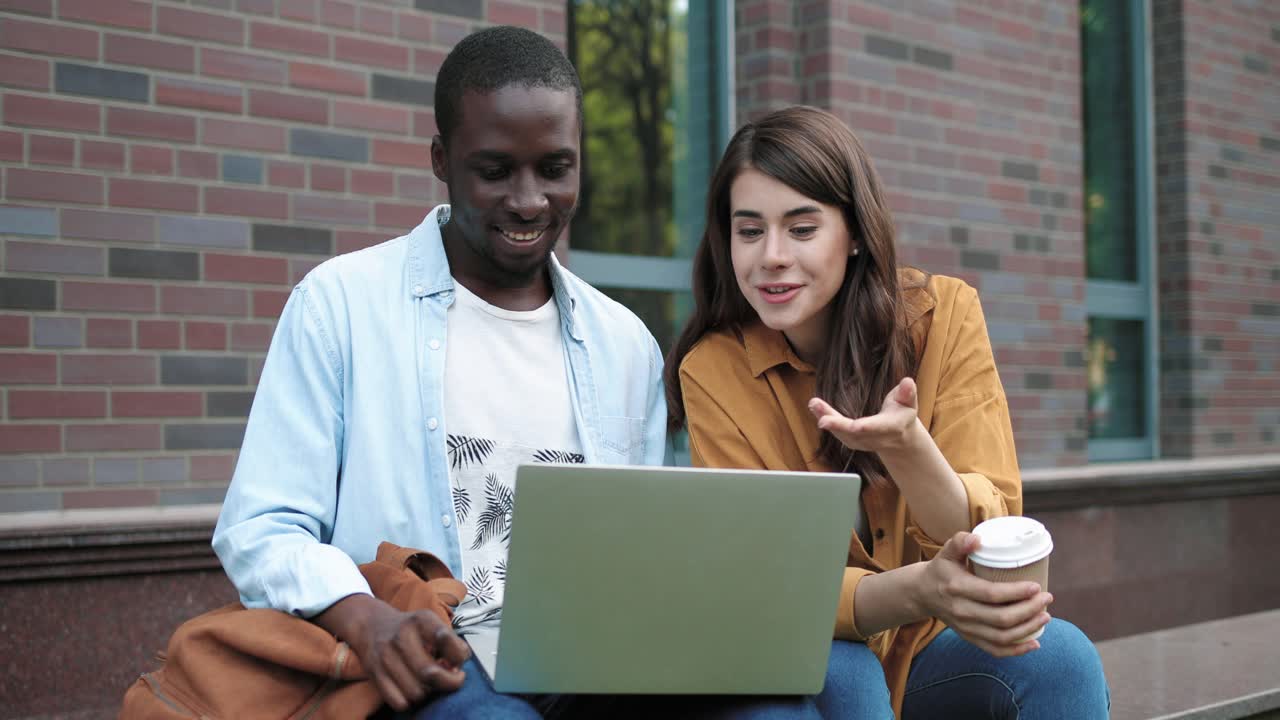 cámara haciendo zoom en un hombre afroamericano y una mujer caucásica sentados en la calle y haciendo una videollamada en una laptop cerca de la universidad
