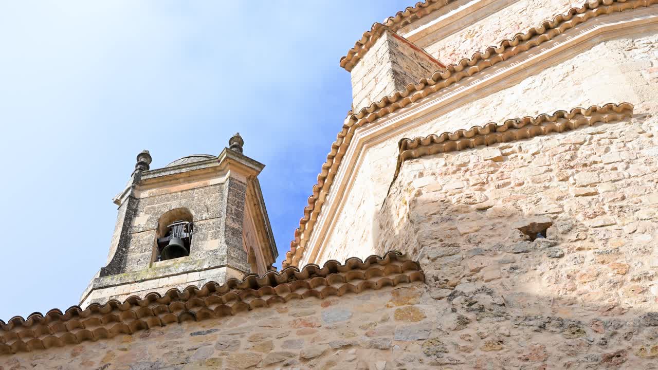 A low-angle, looking-up shot captures the historic church architecture of Cuenca, Spain, detailing its stone walls, terracotta roof, and traditional bell tower