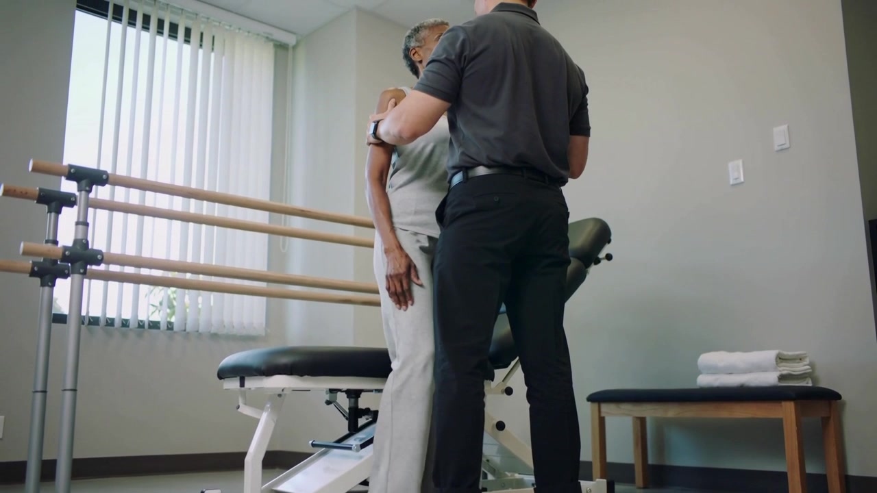 Hände platzierend, Therapeut unterstützt Patienten beim Aufstehen aus dem Sitzen in der Reha mit Gehbarren für das Gleichgewicht