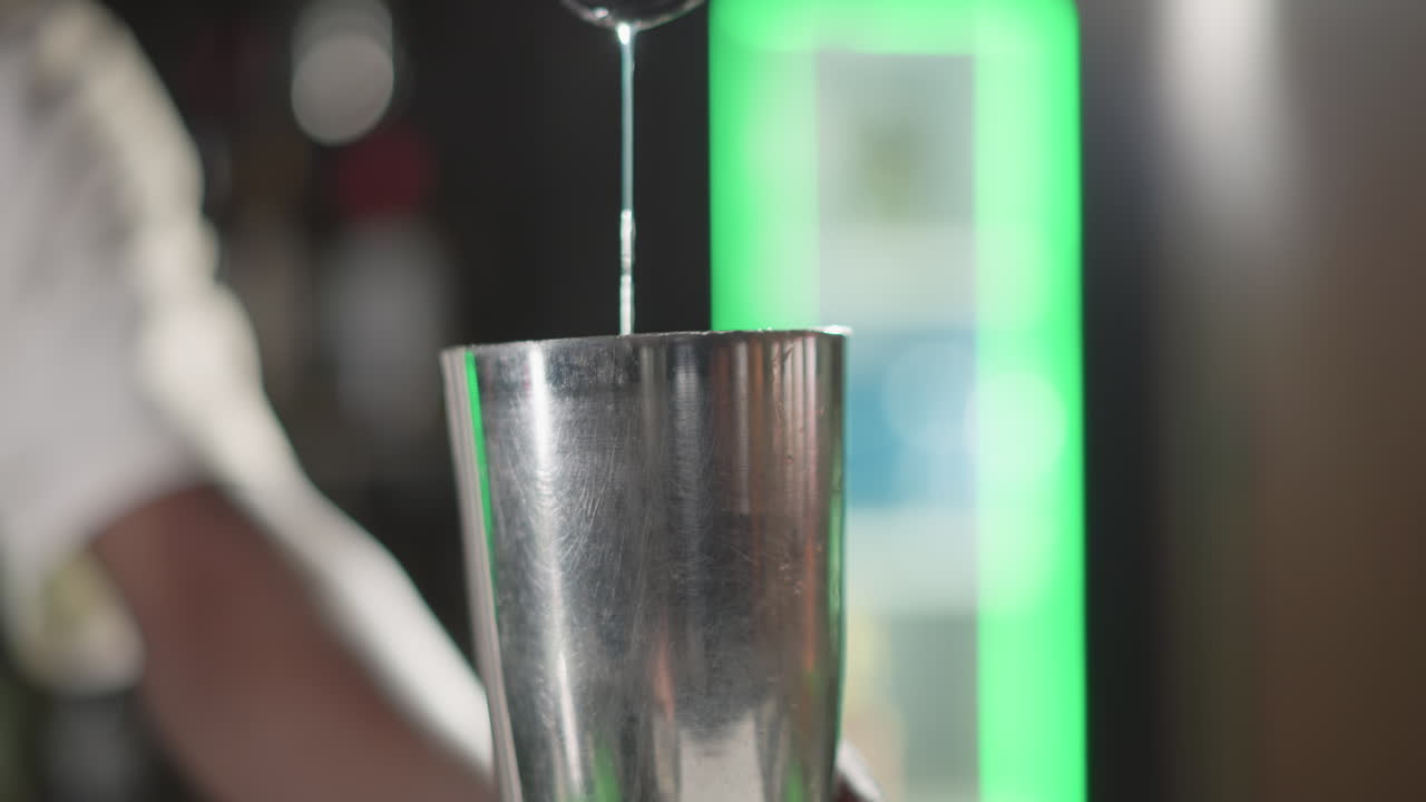 Bartender pouring liquid from jigger into cocktail shaker, focused on mixing ingredients in modern bar with green neon lighting, showcasing bartending skills and precision