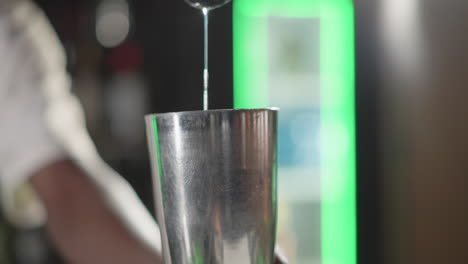 bartender pouring liquid from jigger into cocktail shaker, focused on mixing ingredients in modern bar with green neon lighting, showcasing bartending skills and precision