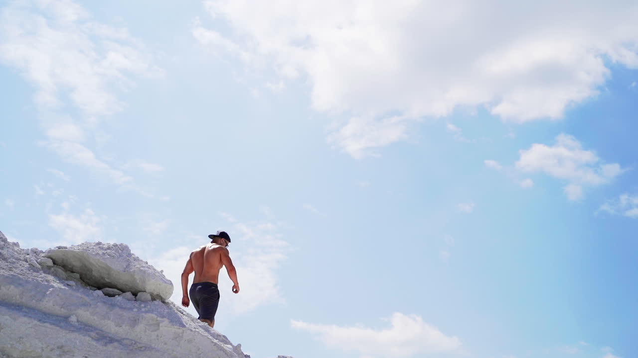 Shirtless man going down the hill. Back view of muscular sportsman in the mountains under blue sky. View from below. Slow motion.
