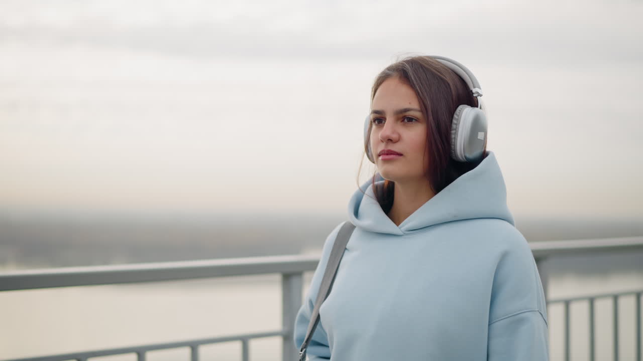Primo piano di una giovane donna sicura di sé che cammina con le cuffie in testa vicino a una ringhiera di ferro con una vista sfocata del fiume sullo sfondo, una passeggiata calma ed elegante in un ambiente urbano all'aperto
