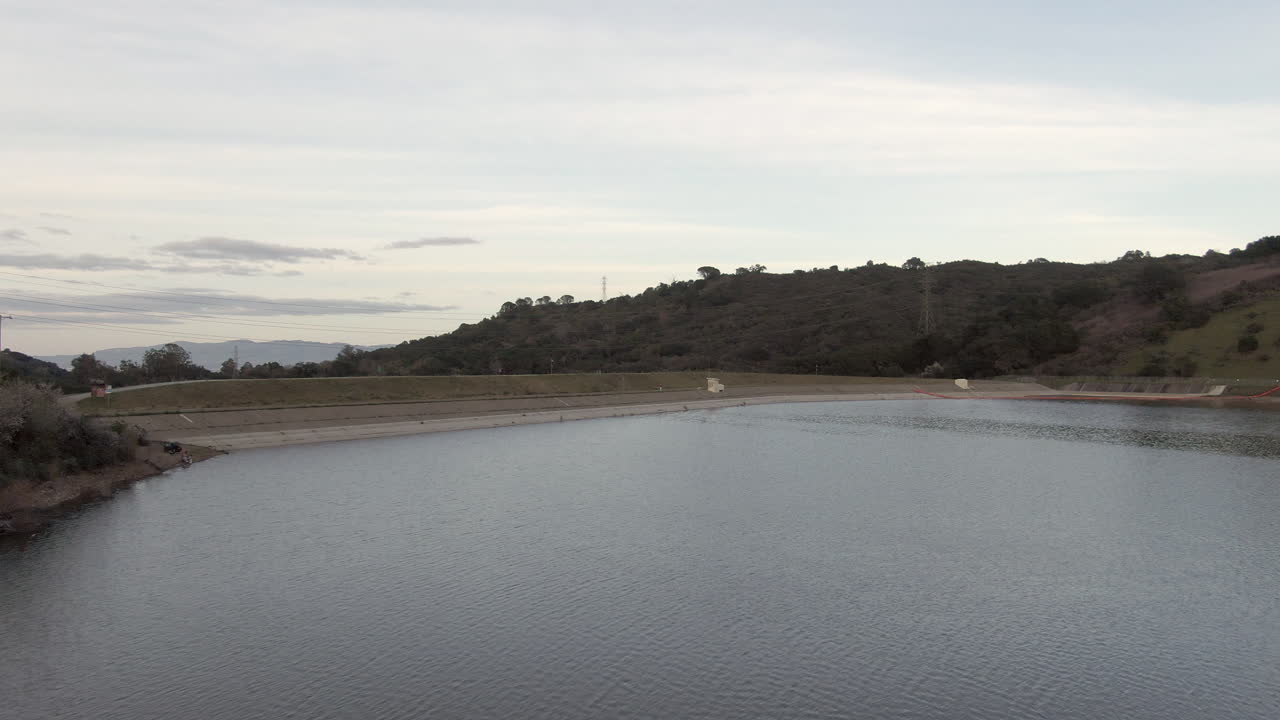Cinematisk sidelæns tracking-optagelse, der glider hen over det dybblå vand i Stevens Creek Reservoir. Det vidtstrakte Silicon Valley-landskab og Cupertinos tech-knudepunkt er synligt i baggrunden under en diset
