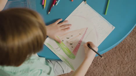 top view of little boy sitting at desk and drawing on paper with colored pencil