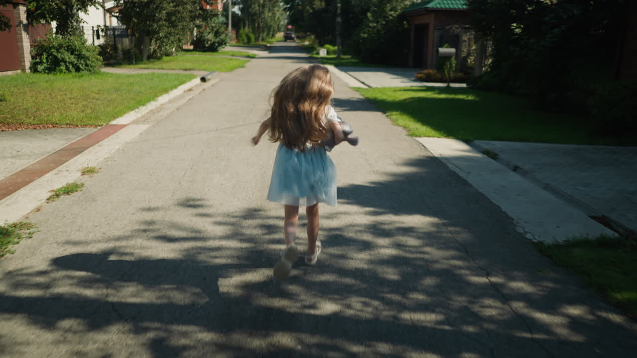 Back view of young girl running down cracked sunny street with long hair swaying in breeze through green suburban neighborhood casting calm soft shadows and motion blur on pavement quietly