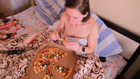 the girl lies in bed covered with blankets and eats delicious pizza with pleasure.