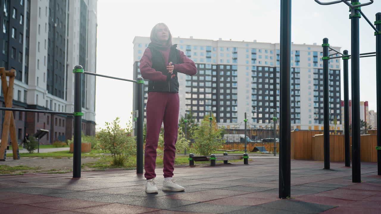 Fitness enthusiast with focused expression standing beside outdoor exercise bars under bright sun rolling hands then neck before workout routine in urban park surrounded by modern apartment buildings