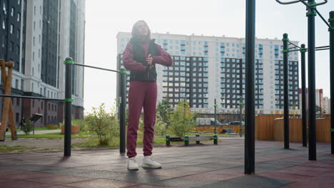 fitness enthusiast with focused expression standing beside outdoor exercise bars under bright sun rolling hands then neck before workout routine in urban park surrounded by modern apartment buildings