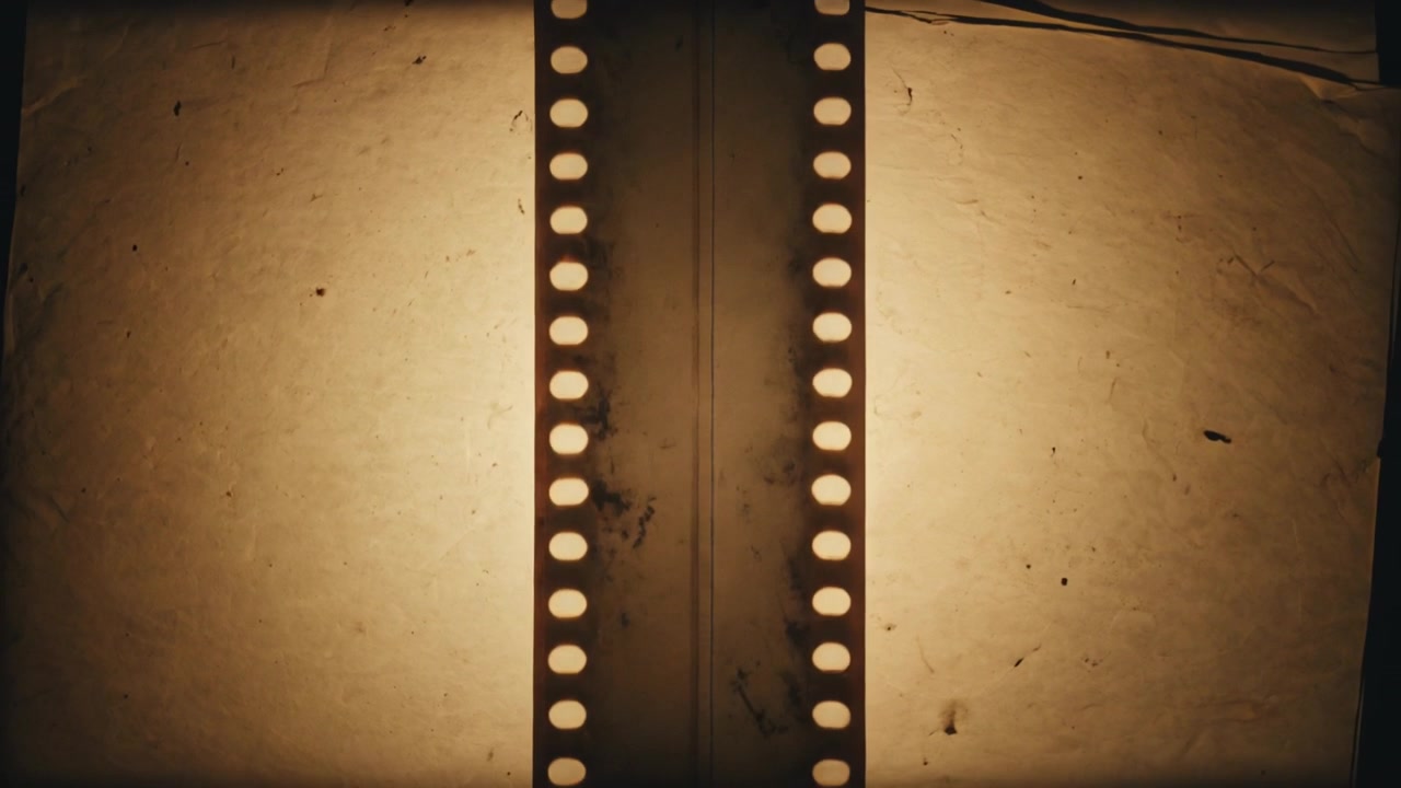 Tira de película de 35mm avanzando por un mecanismo de transporte, moviéndose sobre una caja de luz cálida para escanear, revelando polvo y un cable.