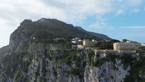 Sunlit-coastal-buildings-on-cliffside-with-lush-greenery,-clear-sky,-daytime,-aerial-shot