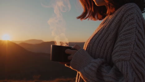 person holding a steaming mug in mountains at sunset