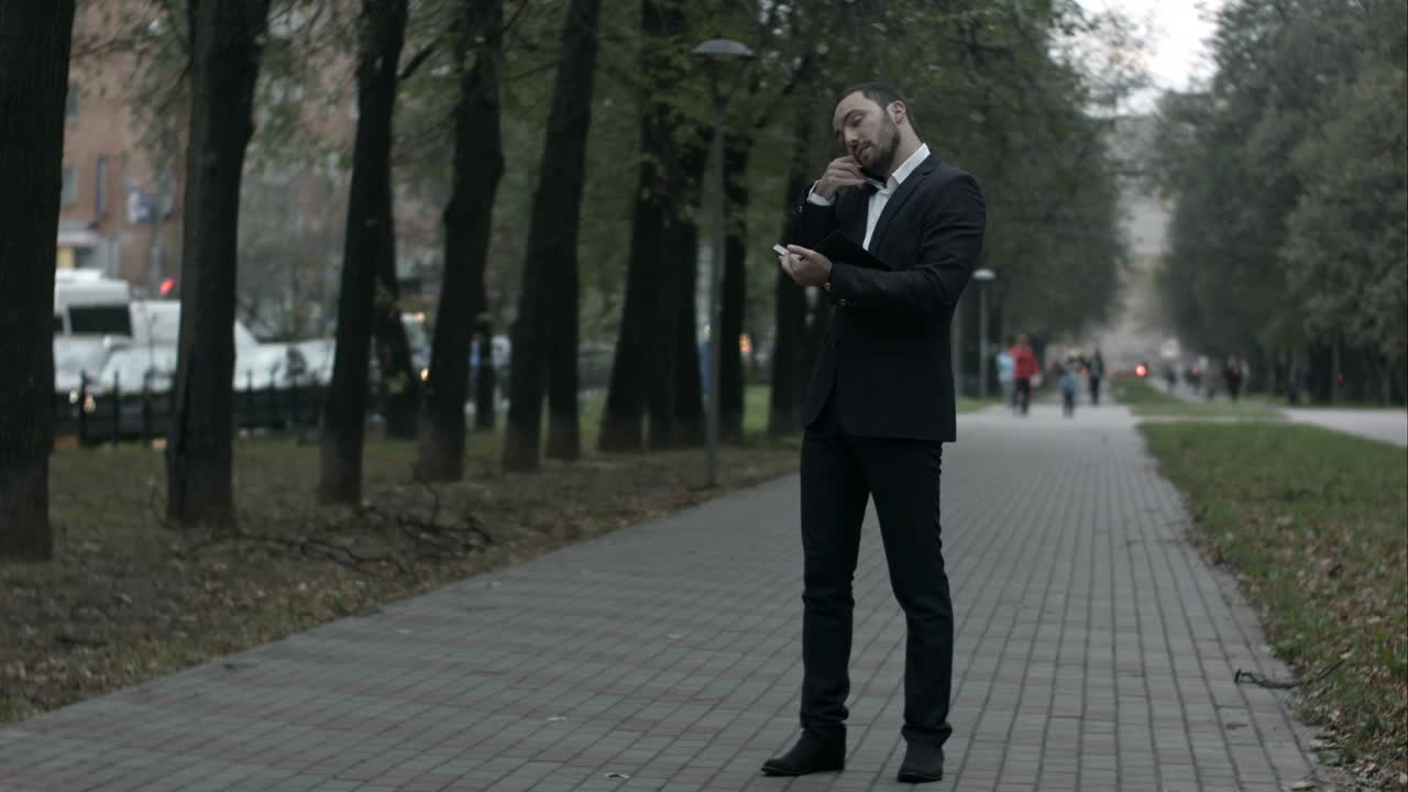 бородатый бизнесмен в парковой аллее разговаривает по телефону и пишет в дневнике, работает с записками в зеленом парке