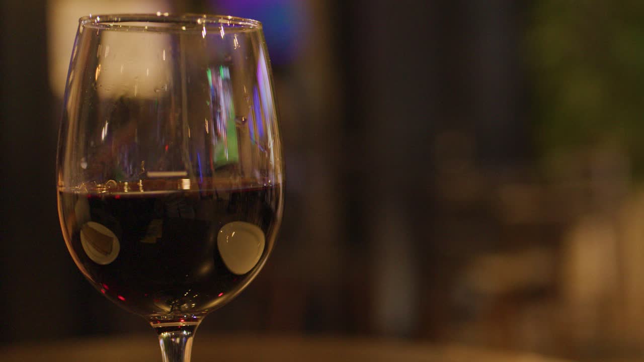Two people clink red wine glasses in warm, softly lit hotel bar with blurred television