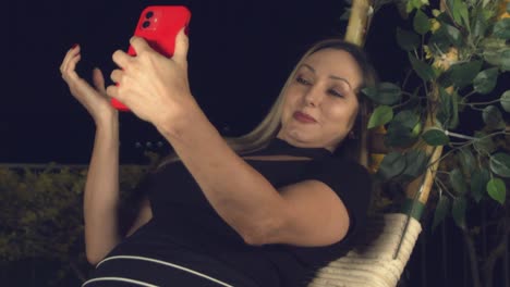 Attractive-woman-leaning-back-on-an-outdoor-swing-at-night-taking-a-selfie