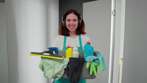 retrato de una confiada bruneta feliz dama de limpieza chica con auriculares inalámbricos negros una camiseta blanca y un delantal gris que camina y sostiene en sus manos un cuenco de plástico gris con detergentes y herramientas de limpia en un apartamento moderno