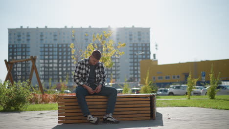 freelancer seated on wooden bench outdoors opens medication pack to take pill surrounded by modern office buildings parked cars bright sunlight and signpost in background under clear sky