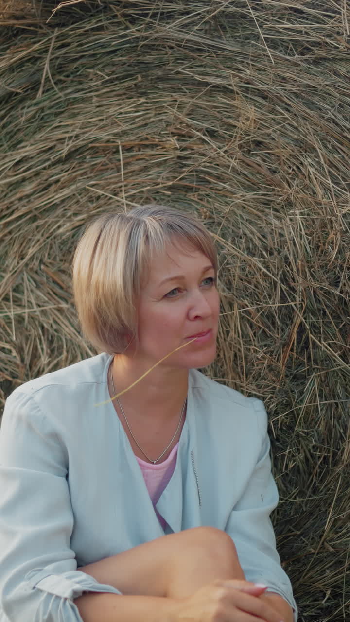 женщина в синем пиджаке сидит на сено, держа сухую палку во рту, позирует с рукой над коленом, мирная сельская обстановка с теплыми естественными тонами захватывает сельскую расслабленность и простоту