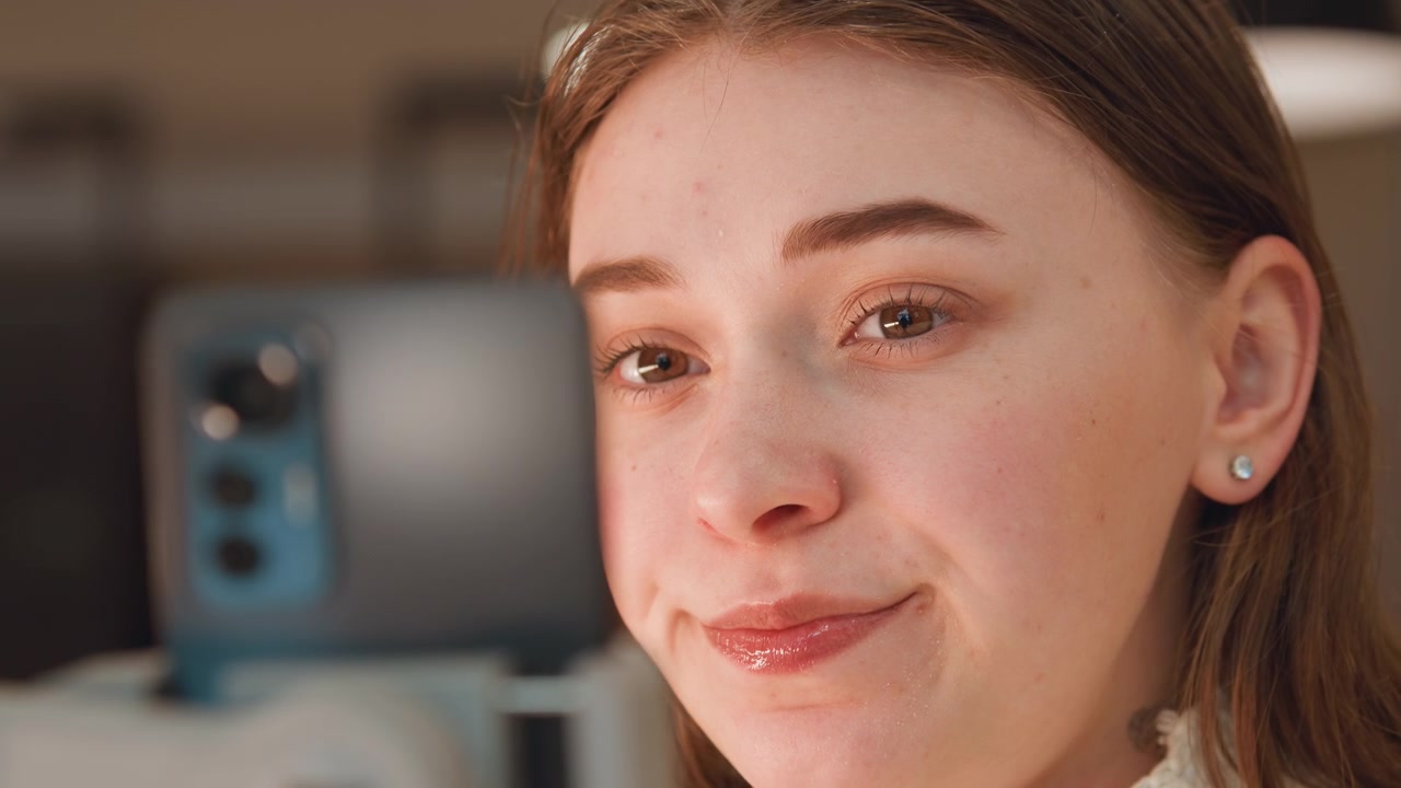 Tight eye-level close-up of young woman interacting with smartphone on mount, progression from focused gaze to gentle smile and relaxed expression indicating pleasant reaction; indoor modern setting