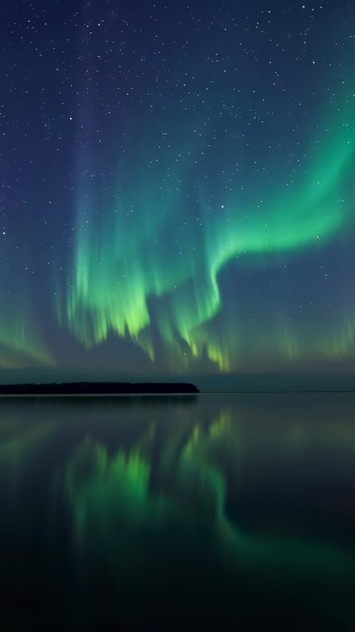 Video verticale: nastri di aurora verde cangiante che brillano e si arricciano su un lago notturno, vento solare scintillante