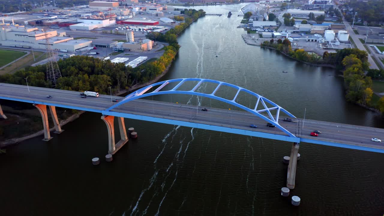 Traffic crosses the arched Leo Frigo Memorial Bridge as it stretches above the calm river lined with industrial buildings and autumn foliage in warm evening light