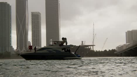 Yacht-in-a-harbor-with-a-man-walking-along-the-edge-of-it,-with-a-city-filled-with-skyscrapers-under-a-cloudy-grey-sky-in-the-background