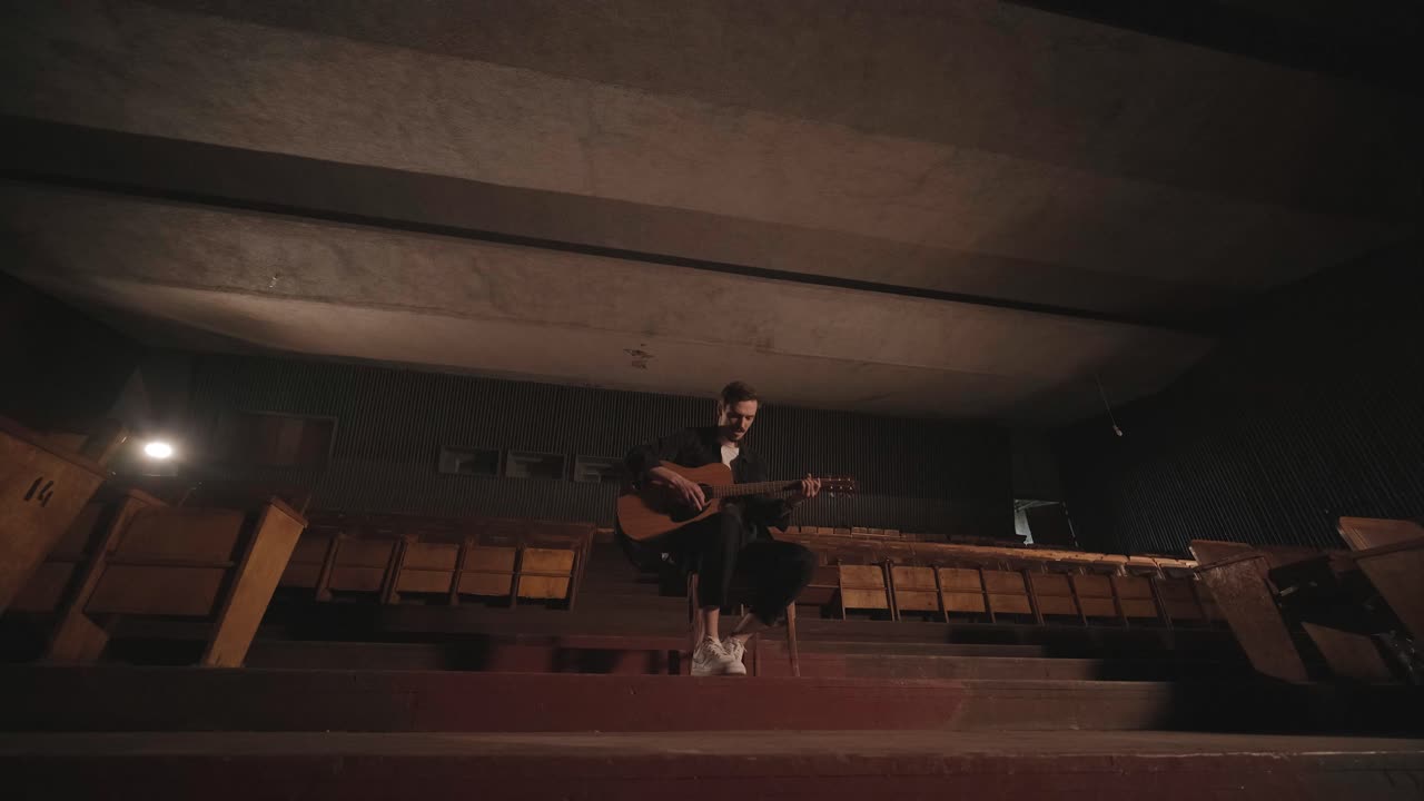 un chico guapo toca una guitarra acústica en un cine abandonado. el músico canta una canción y acompaña en la guitarra