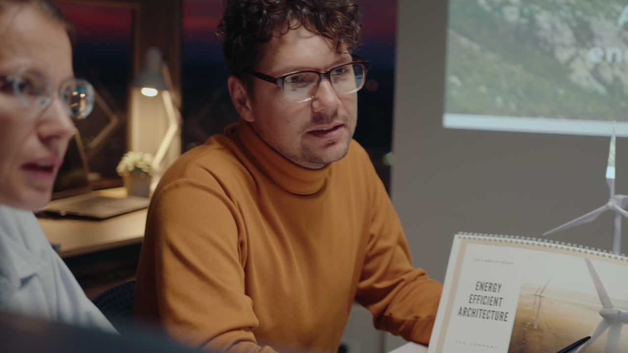 Young man presenting energy efficient architecture project to team of colleagues during business meeting in the office, evening sunset is seen behind windows in the background