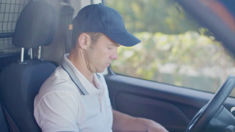 focused courier in cap sitting in car, writing on clipboard and checking the tablet