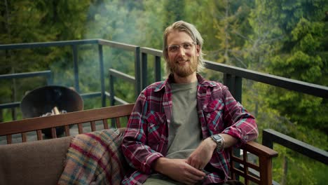 a blond guy in glasses and a red plaid shirt poses while sitting on the couch during a picnic on the balcony of a country house overlooking the mountains and the forest during