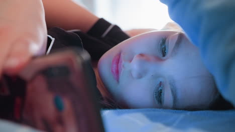 young lady lies on bed under soft blue light while using smartphone and speaking, expression appears thoughtful and engaged, suggesting casual conversation or digital interaction
