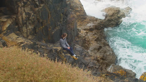 teen lad sitting on coastal cliff edge watching turquoise ocean waves crash over rocks, cornwall, england - static shot