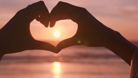 clsoe up woman hands making heart shape gesture holding beautiful sunset flare enjoying romantic vacation on beach