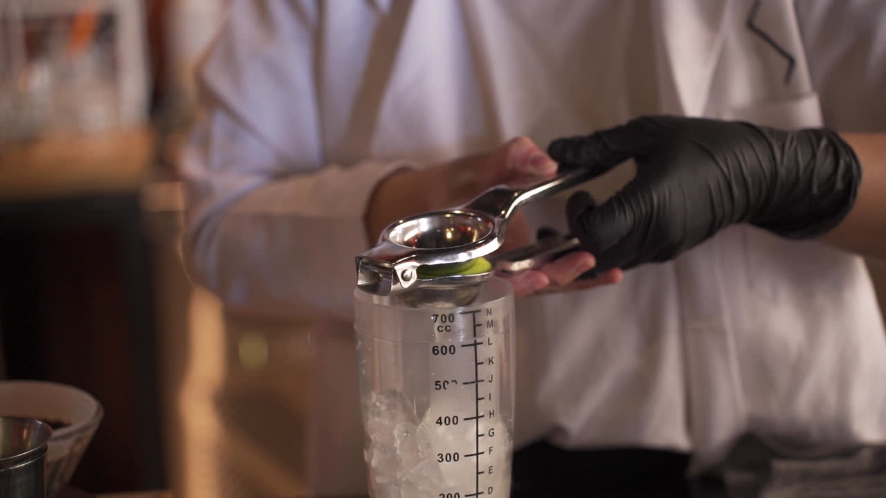 Un barman professionista in uniforme bianca e guanti neri usa uno spremiagrumi di metallo per spremere succo di lime fresco in uno shaker di plastica graduato pieno di cubetti di ghiaccio mentre prepara un cocktail