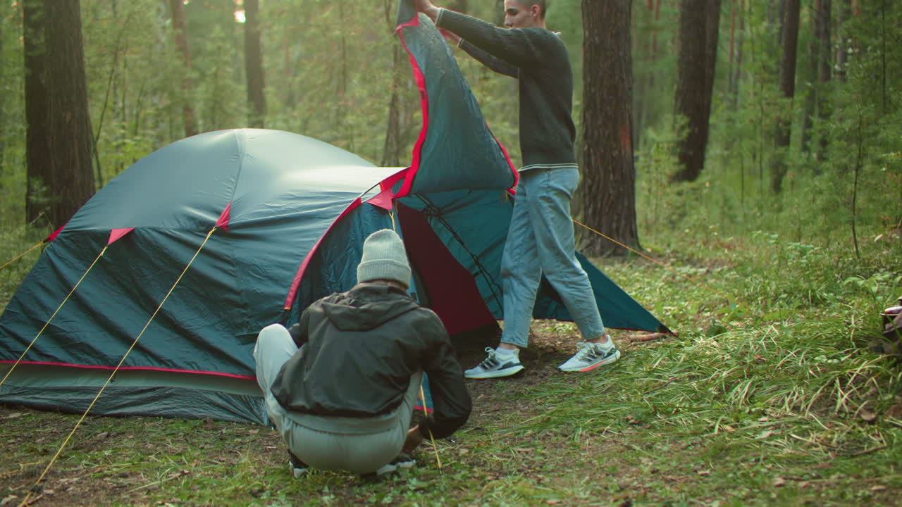 Young man lifts tent cover with admiration while woman squats at entrance holding rope securing peg in dense forest under soft sunlight during camping setup with knit beanie and casual sweater