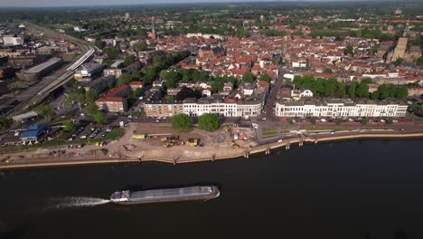 Aerial-following-of-inland-shipping-large-cargo-vessel-leaving-ripple-waves-on-river-IJssel-passing-IJsselkade-boulevard-countenance-cityscape-of-tower-town-Zutphen