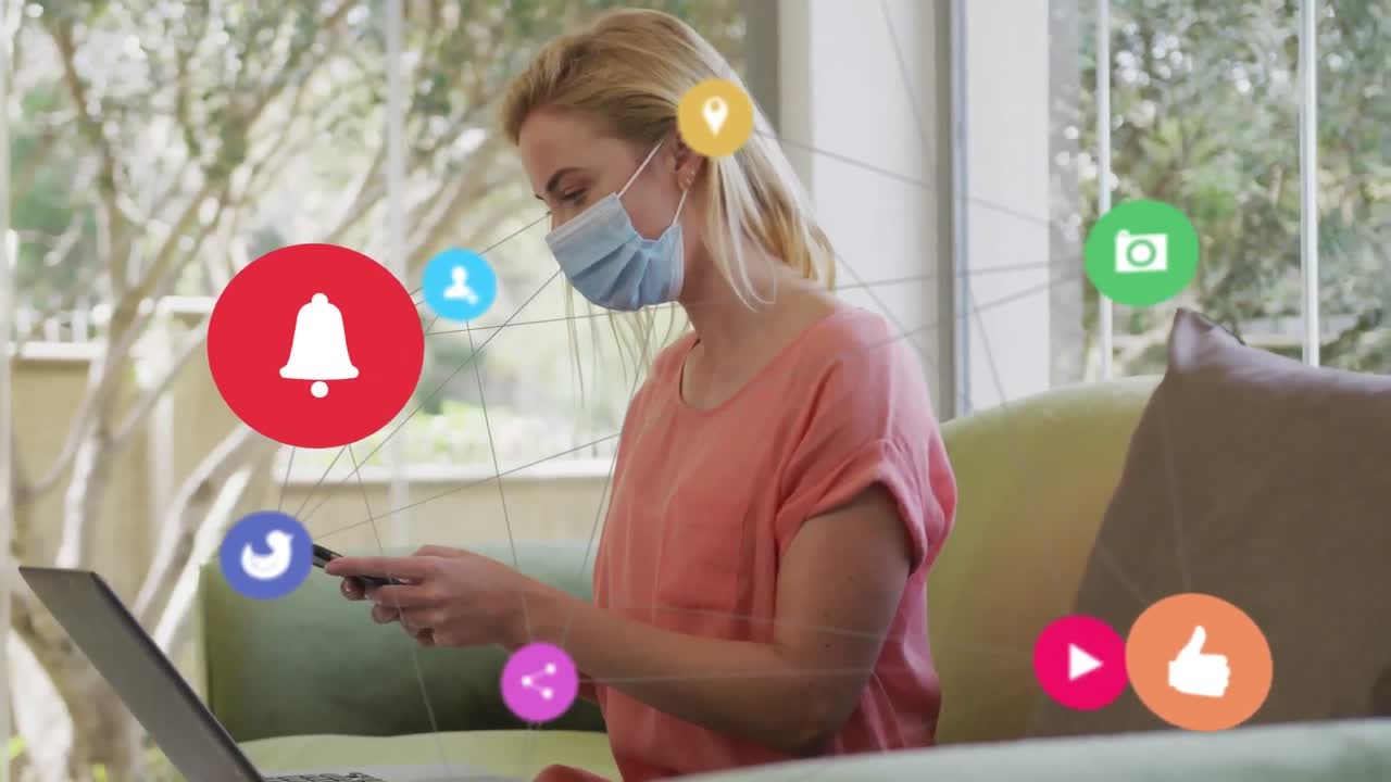 Animation of digital icons over caucasian woman in face mask using smartphone at home