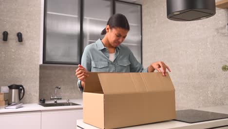 joven afroamericana decepcionada de pie en la cocina con una caja de cartón y sintiéndose molesta