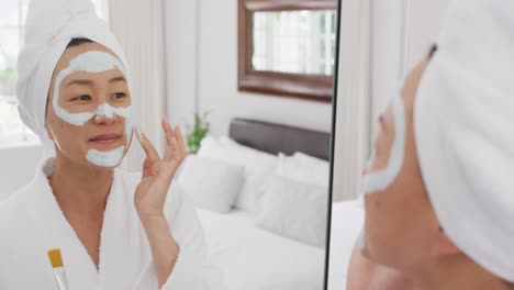 video of happy asian woman in robe with moisturizing mask looking at mirror