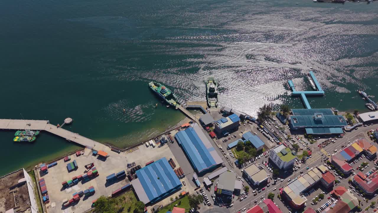 Orbit drone shot of Victoria Jetty on Labuan Island, Malaysia, showing Ro-Ro ship, car carrier, and port buildings under sunlight reflecting on ocean waves. Concept of transport or maritime footage