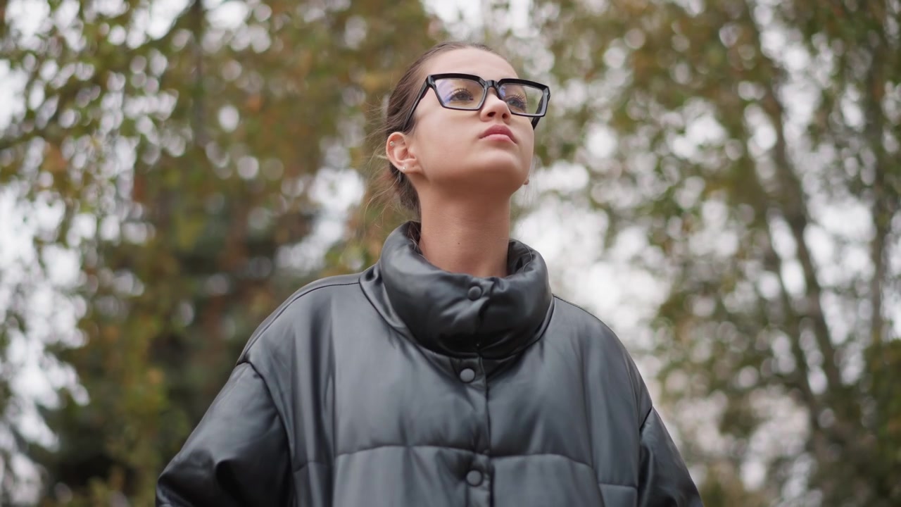 Primo piano di una donna che indossa occhiali neri e una giacca di pelle scura, in piedi all'aperto con alberi sfocati. La sua espressione è calma mentre guarda in alto, catturando la luce naturale sul suo viso
