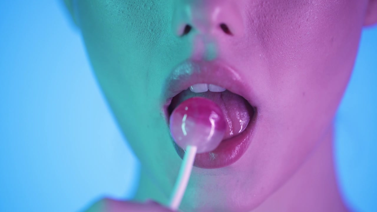 Este cautivador video en cámara lenta 4K presenta un primer plano hipnotizante de una mujer disfrutando de una piruleta rosa redonda sobre un llamativo fondo azul. La escena está bellamente realzada.