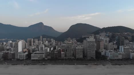 Antena-Retrocediendo-Desde-La-Playa-Vacía-De-Ipanema-Temprano-En-La-Mañana-Revelando-Las-Olas-Del-Océano-En-Primer-Plano-Contra-El-Paisaje-Urbano-Más-Amplio-De-Río-De-Janeiro-Con-La-Montaña-Corcovado-En-El-Fondo