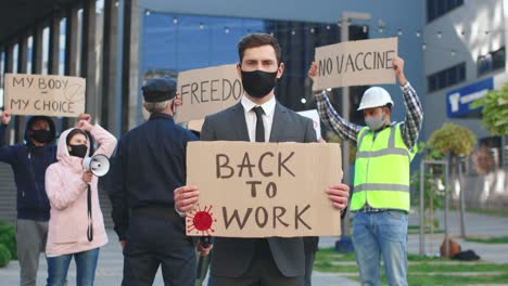 hombre de negocios con máscara facial y traje reteniendo el cartel de "trabajo" y mirando la cámara en una protesta contra covid 19"