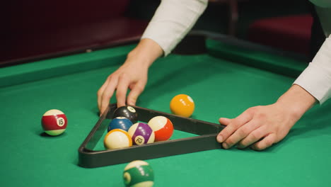 gentleman in white shirt arranging billiard balls using triangle rack on green pool table. hands carefully aligning balls for precise setup. close-up shot capturing focused preparation